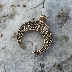 May include: A bronze-colored crescent moon pendant with intricate scrollwork and a small loop at the top. The pendant has a detailed, ornate design with a textured surface and a polished finish. The crescent moon is a symbol of femininity and change.
