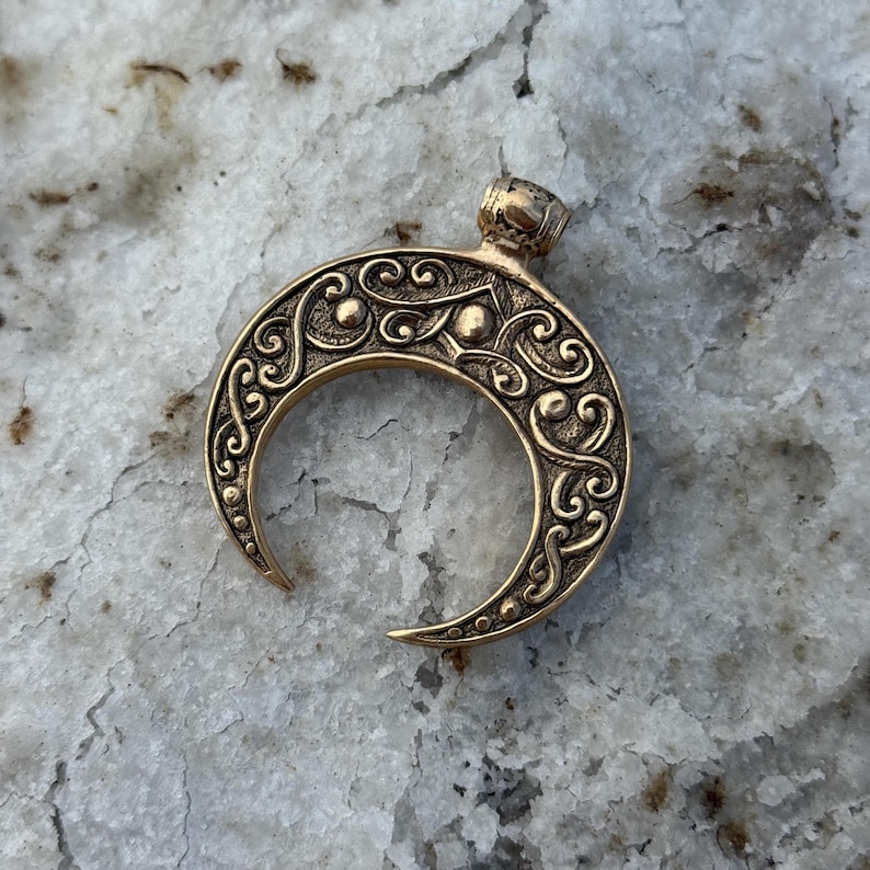 May include: A bronze-colored crescent moon pendant with intricate scrollwork and a small loop at the top. The pendant has a detailed, ornate design with a textured surface and a polished finish. The crescent moon is a symbol of femininity and change.