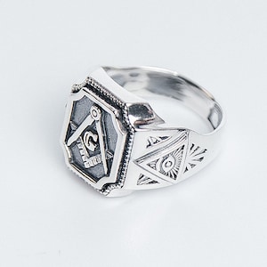 K&ouml;nnte beinhalten: Ein silberner Freimaurer-Ring mit einem Quadrat- und Zirkel-Design und einem allsehenden Auge auf dem Band.