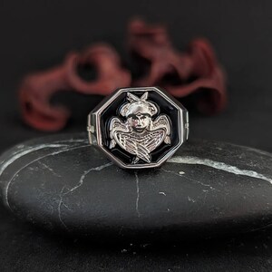 K&ouml;nnte beinhalten: Silberfarbener Ring mit achteckiger schwarzer Fassung, die ein detailliertes Engeldesign zeigt. Der Engel hat Fl&uuml;gel und einen ruhigen Ausdruck. Der Ring liegt auf einer dunklen, strukturierten Oberfl&auml;che, mit verschwommenen roten Dekorationselementen im Hintergrund.