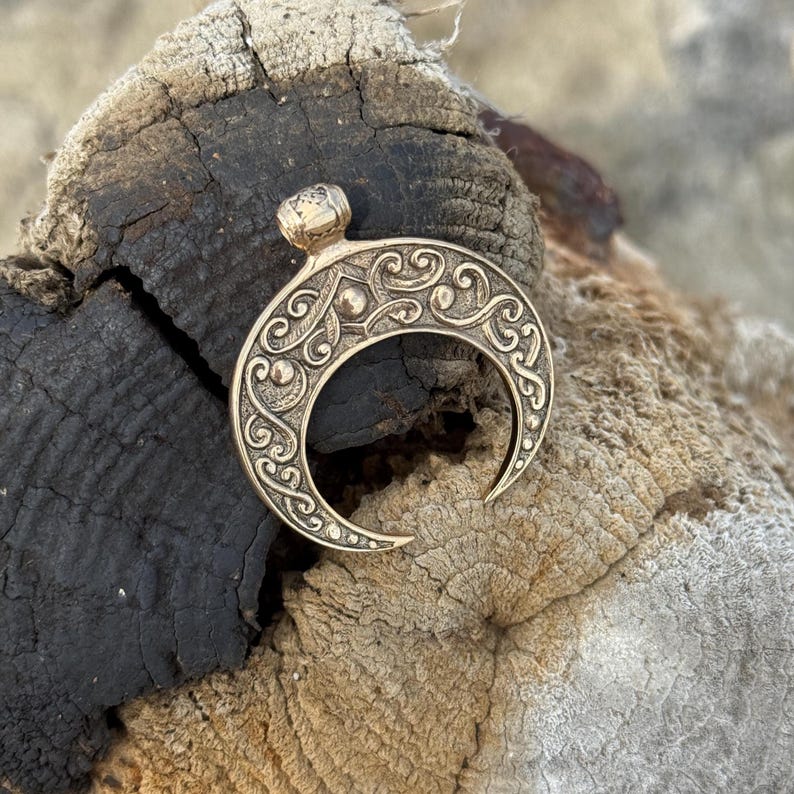 May include: A gold-colored crescent moon pendant with intricate scrollwork and a small, rounded top. The pendant is likely made of metal and features a detailed, embossed design. The background is a weathered, dark wood surface.