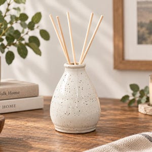 May include: A white ceramic reed diffuser bottle with a speckled black pattern and a tapered neck. Four light-colored wooden reeds are inserted into the opening of the bottle, extending upwards from the center of the vessel.