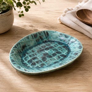 May include: An oval ceramic dish featuring a textured, grid-like pattern with a glossy glaze in shades of turquoise, teal, and dark blue. The surface has a reactive finish with darker speckles concentrated within the recessed grid sections.