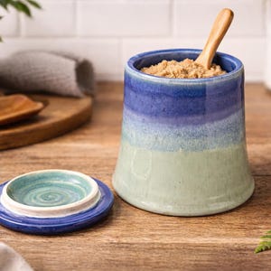 May include: A ceramic sugar bowl with a blue and green ombre design, filled with brown sugar and a wooden spoon. A matching lid sits beside it. The kitchenware is on a wooden surface.