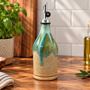 May include: A ceramic oil dispenser bottle with a speckled beige base and a glossy teal glaze dripping down from the neck. The bottle features a stainless steel pour spout with a black rubber stopper, designed for controlled liquid dispensing.