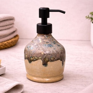 May include: A ceramic soap dispenser with a black pump. The dispenser has a beige base with a textured, glazed finish in shades of brown, blue, and green. The dispenser is on a light-colored surface.