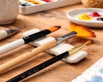 May include: A collection of artist's paintbrushes, including a fan brush, resting on white ceramic brush rests. A palette of watercolors and a small ceramic cup are in the background, suggesting a painting setup. The wooden surface adds a natural touch.