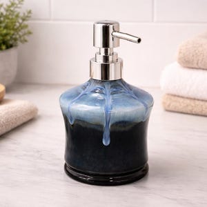 May include: A ceramic soap dispenser with a blue and black glaze, topped with a silver pump. The dispenser has a rounded shape and sits on a marble surface. Soft towels are stacked in the background.