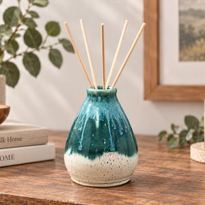 May include: A ceramic reed diffuser bottle with a tapered neck and a rounded base. The upper portion features a glossy, deep teal glaze that drips down into a speckled, off-white matte base. Five light-colored wooden reeds extend from the opening.
