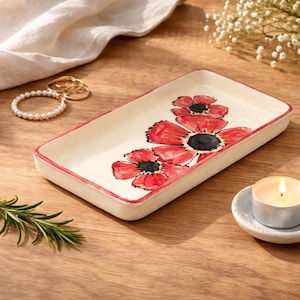 May include: A rectangular ceramic tray with an off-white base and a hand-painted red poppy flower design. The tray features a thin red border along the edges, providing a clean and simple look for holding small items like jewelry or trinkets.