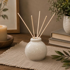May include: A small, speckled ceramic diffuser vase with wooden reeds. A lit candle in a wooden bowl and a framed botanical print are in the background. A potted plant and books are also visible, creating a cozy, neutral-toned aesthetic.