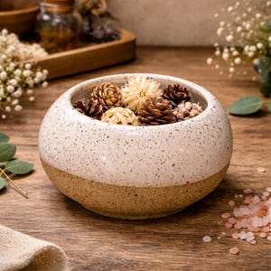 May include: A speckled, off-white ceramic bowl with a brown base, filled with dried pine cones and decorative spheres. The bowl sits on a wooden surface, suggesting a rustic or natural theme.