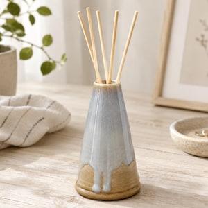 May include: A conical ceramic reed diffuser with a reactive glaze featuring a smooth transition from a light blue-grey top to a tan base. The vessel has a wide bottom and a narrow neck, containing five thin, light-coloured wooden diffuser reeds.