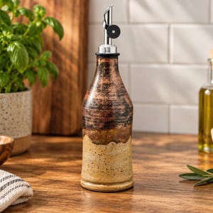 May include: A ceramic oil dispenser bottle with a two-tone finish, featuring a speckled beige base and a dark, textured brown upper section. The bottle is topped with a silver-colored metal pour spout that includes a small black circular stopper.