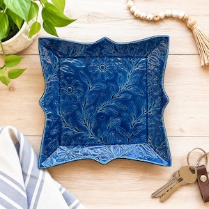 May include: A square, deep blue ceramic dish with scalloped edges and a raised, embossed floral and leaf pattern across the entire surface. The glaze has a glossy finish, highlighting the intricate botanical details carved into the clay.