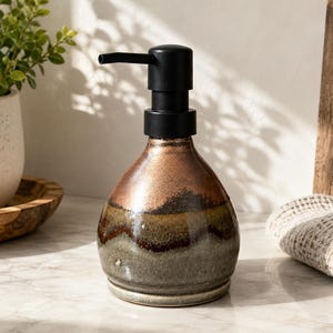 May include: A ceramic soap dispenser with a rounded base featuring a reactive glaze in shades of copper, brown, and sage green. The dispenser is topped with a matte black plastic pump mechanism that extends horizontally from the top center.