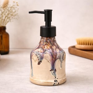 May include: A ceramic soap dispenser with a black pump. The dispenser has a beige base with dripped glaze in shades of purple, brown, and white. The dispenser is on a light-colored surface, with a stack of towels and a vase in the background.