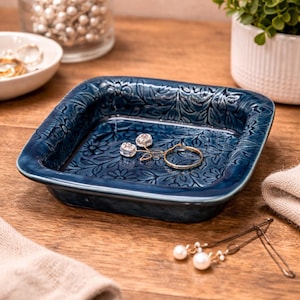 May include: A square, navy blue ceramic jewelry dish with a floral embossed design. The dish holds a gold ring and two clear, sparkling earrings. Other jewelry and accessories are visible in the background.