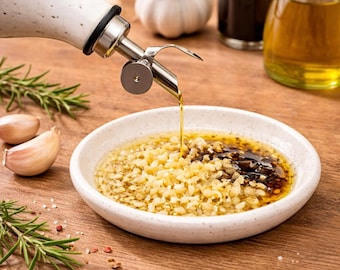 May include: A ceramic oil dispenser with a stainless steel spout pours oil into a white bowl filled with minced garlic and other ingredients. Garlic cloves, rosemary sprigs, and other bottles are in the background on a wooden surface.