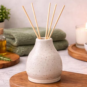 May include: A speckled ceramic essential oil diffuser with wooden reeds. The image also features a lit candle, a bowl of potpourri, a sliced orange, and a small bottle of oil, all arranged on wooden trays. Green towels and a potted plant are in the background.
