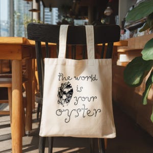 Peut inclure: Sac fourre-tout crème avec l'inscription "the world is your oyster" en script noir. Le sac présente un graphisme noir et blanc d'une coquille d'huître. Le sac est accroché au dossier d'une chaise en bois noir.