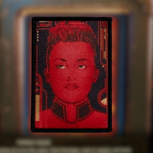 May include: An embroidered patch featuring a portrait of a person with a serious expression. The person is wearing a dark blue and red uniform with gold trim. The background is dark blue with a pattern of lines and dots.