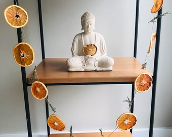 May include: A decorative garland made of dried orange slices and sprigs of green rosemary strung together on a thin cord, draped in front of a white Buddha statue sitting in a meditative pose on a wooden shelf with a black metal frame.
