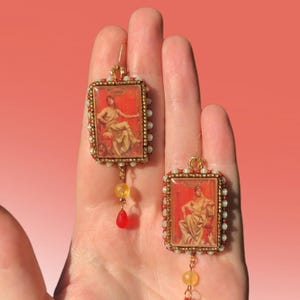 Op de afbeelding: Een paar gouden oorbellen met een rechthoekig frame in rood en goud met een vintage afbeelding van een vrouw. Elke oorbel heeft een gele en rode kralenhanger.