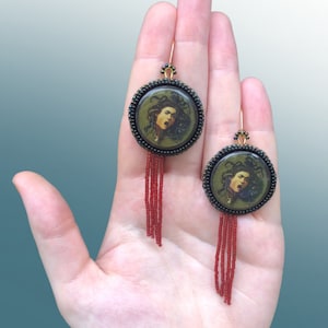 May include: A pair of earrings with a round, green and black beaded frame featuring a printed image of Medusa's head. The earrings have red beaded tassels.