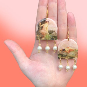 May include: A pair of gold hoop earrings with a vintage painting of a woman in a landscape scene. Each earring has three white pearl beads hanging from the bottom.