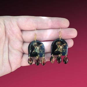 Puede incluir: Un par de pendientes de aro dorados con dijes en forma de arco negro y azul que representan una imagen vintage de una mujer. Los dijes están adornados con tres cuentas en forma de lágrima rojas y doradas.