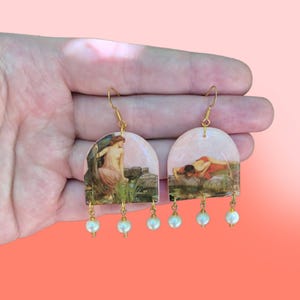 Puede incluir: Un par de pendientes de aro dorados con una pintura vintage de una mujer en un estanque. Los pendientes tienen un colgante de perla.