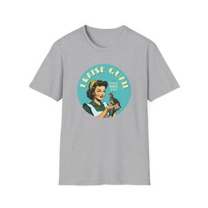 Puede incluir: Una camiseta gris con un diseño de estilo retro. El diseño es un círculo turquesa con las palabras "I RAISE QUAIL" y "BECAUSE MURDER IS BAD". Dentro del círculo, una mujer sostiene una codorniz. La mujer lleva un delantal verde y un pañuelo amarillo.