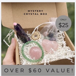 May include: A cardboard box labeled "MYSTERY CRYSTAL BOX" filled with crystals and a bracelet. The box contains a large amethyst crystal, a rose quartz heart, a green beaded bracelet, and other crystals. The text "OVER $60 VALUE!" is at the bottom.