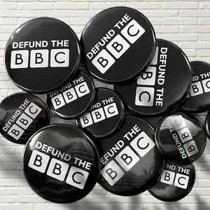 May include: A collection of black and white round buttons with the text "DEFUND THE BBC" printed on them. The buttons are arranged in a pile.