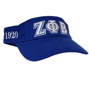May include: A royal blue visor with white embroidered text. The text on the visor reads "ZΦB" above the brim and "1920" on the side. The visor is made of a sturdy fabric and has a curved brim.