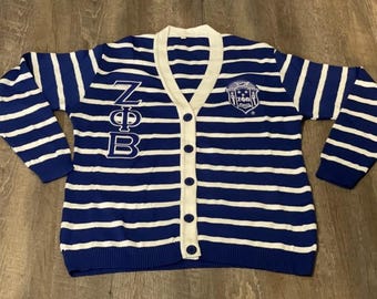 Op de afbeelding: Een blauw met wit gestreept vest met V-hals, voorzien van verticaal geborduurde Griekse letters ZΦB aan de rechterkant en een cirkelvormig ZΦB-embleem op de linkerborst. Het kledingstuk heeft een witte knoopsluiting en lange mouwen.