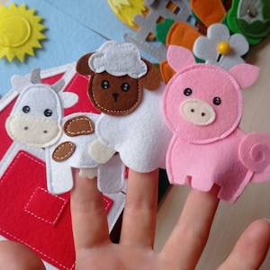 Puede incluir: Un conjunto de marionetas de dedo con temática de granja hechas de fieltro, que incluyen una vaca, una oveja y un cerdo. Las marionetas son blancas, rosas y marrones con ojos de botón negros. Elementos adicionales de granja de fieltro.