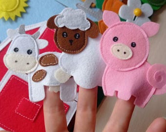 May include: A felt farm animal finger puppet set featuring a cow, sheep, and pig. The puppets are white, pink, and brown with black eyes. The background includes a felt farm scene with a red barn, blue sky, and various felt farm elements.