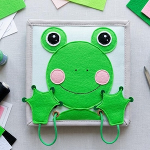 May include: A square felt craft project featuring a green frog applique with large black eyes, pink cheeks, and two green webbed hands attached by green cords. The project is surrounded by green, pink, and black felt sheets, thread spools, and scissors.