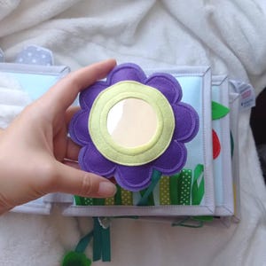 May include: A soft fabric activity book featuring a purple felt flower with a central circular mirror. The page has light blue fabric, green ribbon grass, and stitched details. The book is constructed with light gray fabric binding and edges.