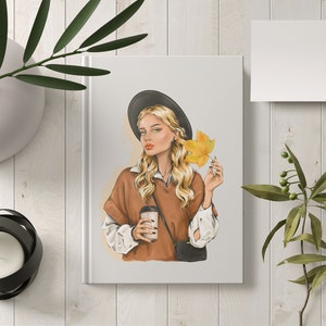 Pode incluir: Uma mulher com cabelo loiro usando um chap&eacute;u preto e um su&eacute;ter marrom segura uma x&iacute;cara de caf&eacute; e uma folha amarela.