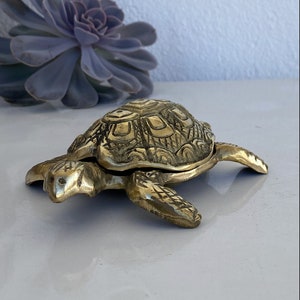 以下が含まれることがあります： 詳細な甲羅のデザインが施された金色の真鍮製の亀の置物。亀は白い表面の上に置かれています。