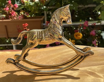 Puede incluir: Una figurita de caballo de balancín de latón con un diseño detallado. El caballo está en una pose de galope con la cabeza girada hacia un lado. La base de balancín está hecha de dos piezas curvas de latón.
