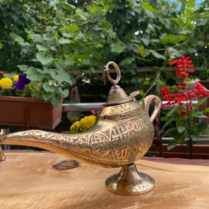 May include: A gold genie lamp with intricate engraved designs and Arabic script. The lamp has a spout, a lid, and a round base. It is sitting on a wooden surface.