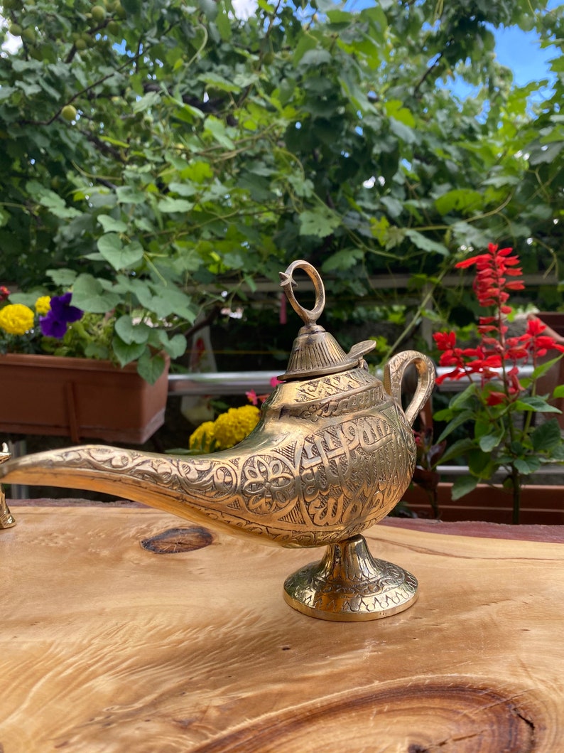 May include: A gold genie lamp with intricate engraved designs and Arabic script. The lamp has a spout, a lid, and a round base. It is sitting on a wooden surface.