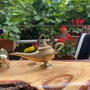 May include: A gold genie lamp with intricate details sits on a wooden table. The lamp has a spout and a handle, and the top is decorated with a ring.