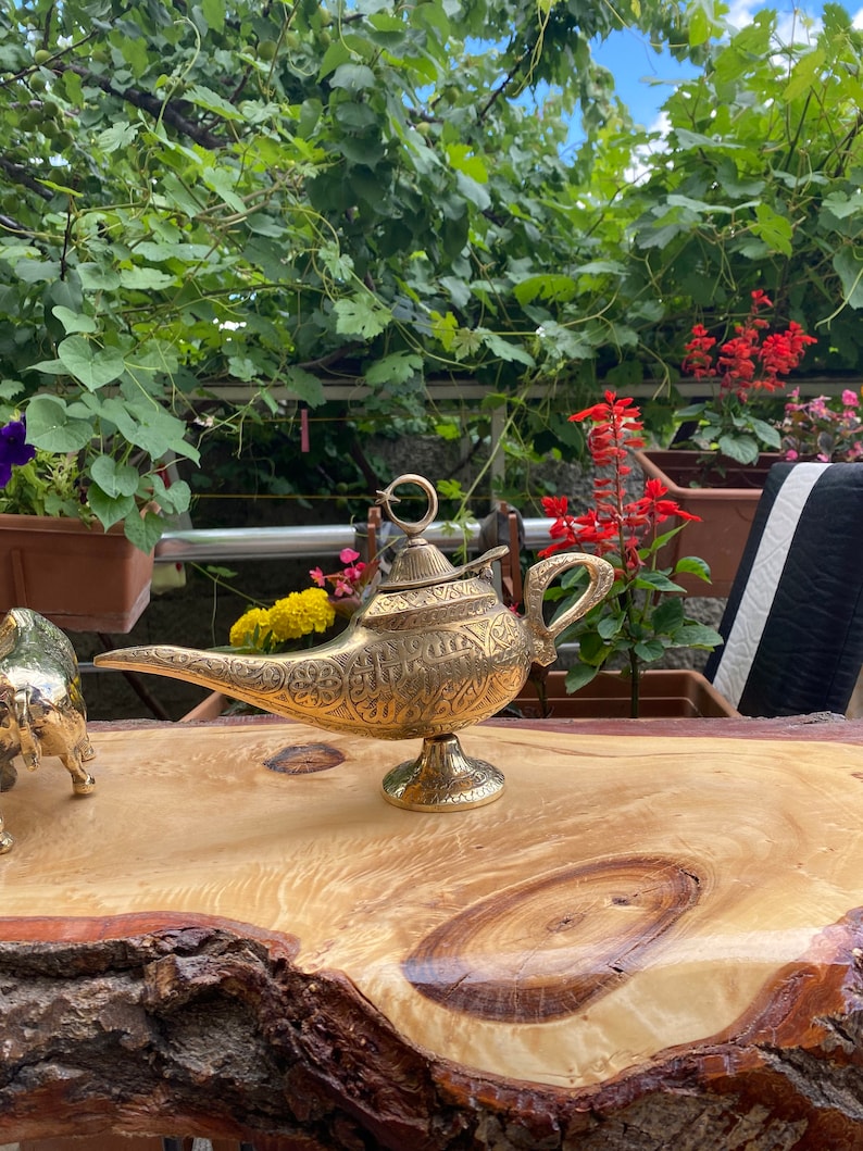 May include: A gold genie lamp with intricate details sits on a wooden table. The lamp has a spout and a handle, and the top is decorated with a ring.