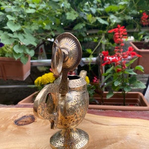 May include: A gold-colored metal teapot with intricate floral designs. The teapot has a round body and a spout with a curved handle. The teapot is sitting on a wooden surface.