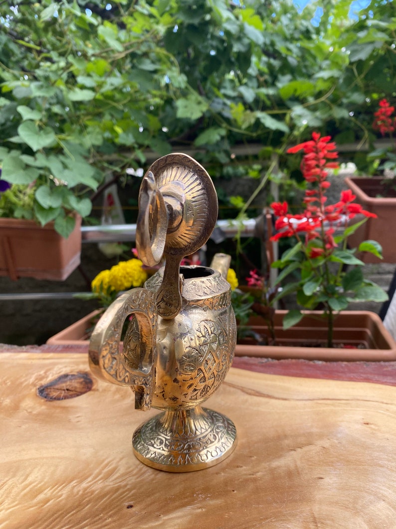 May include: A gold-colored metal teapot with intricate floral designs. The teapot has a round body and a spout with a curved handle. The teapot is sitting on a wooden surface.